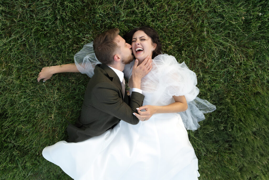 The bride and groom laying in the grass and the groom is kissing the birde on the cheek.